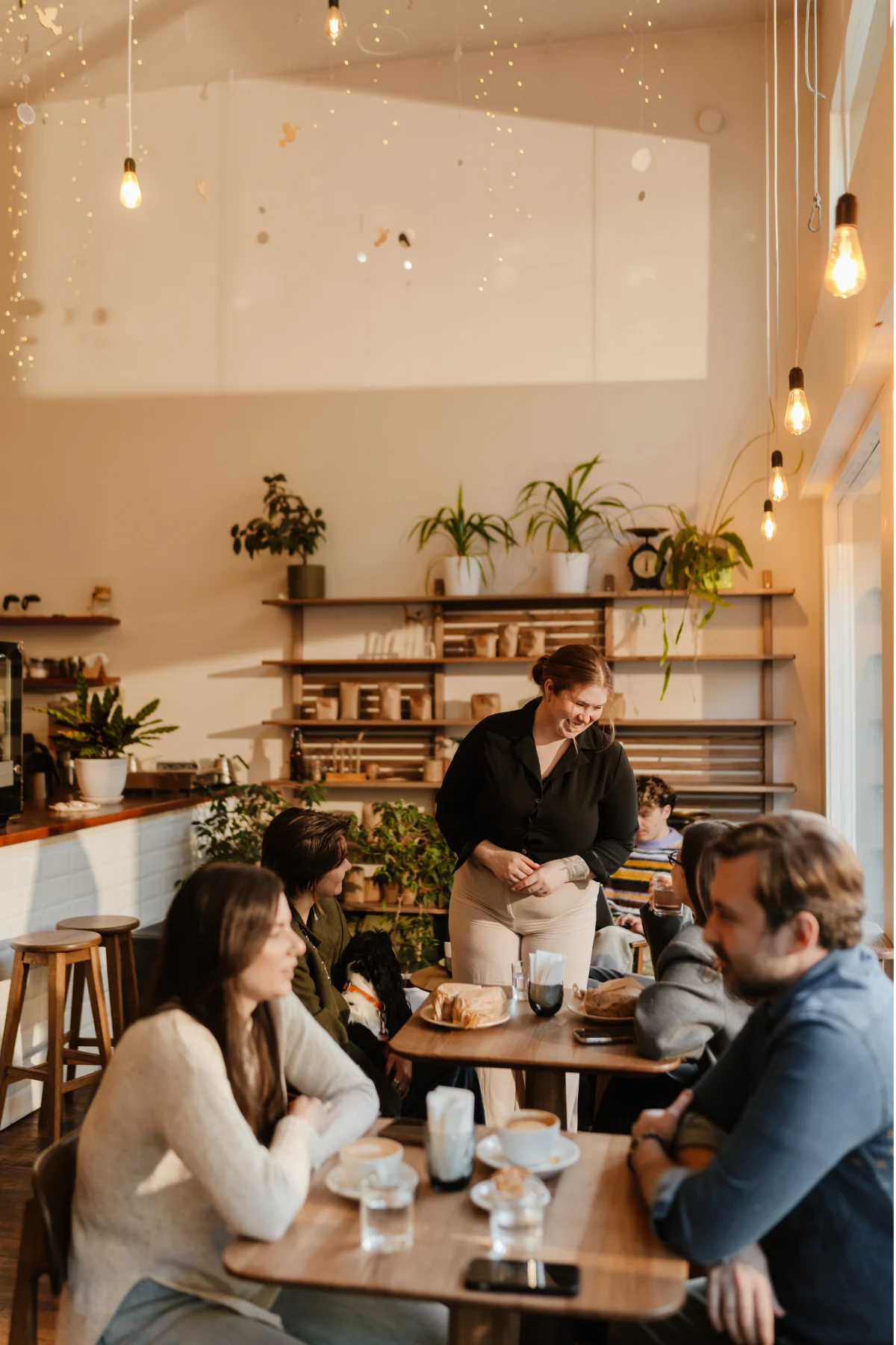 Cozy Café Scene A cozy café interior with several patrons seated at wooden tables. A staff member stands by one table, taking an order. The atmosphere is warm with soft lighting and green plants decorating the space.
