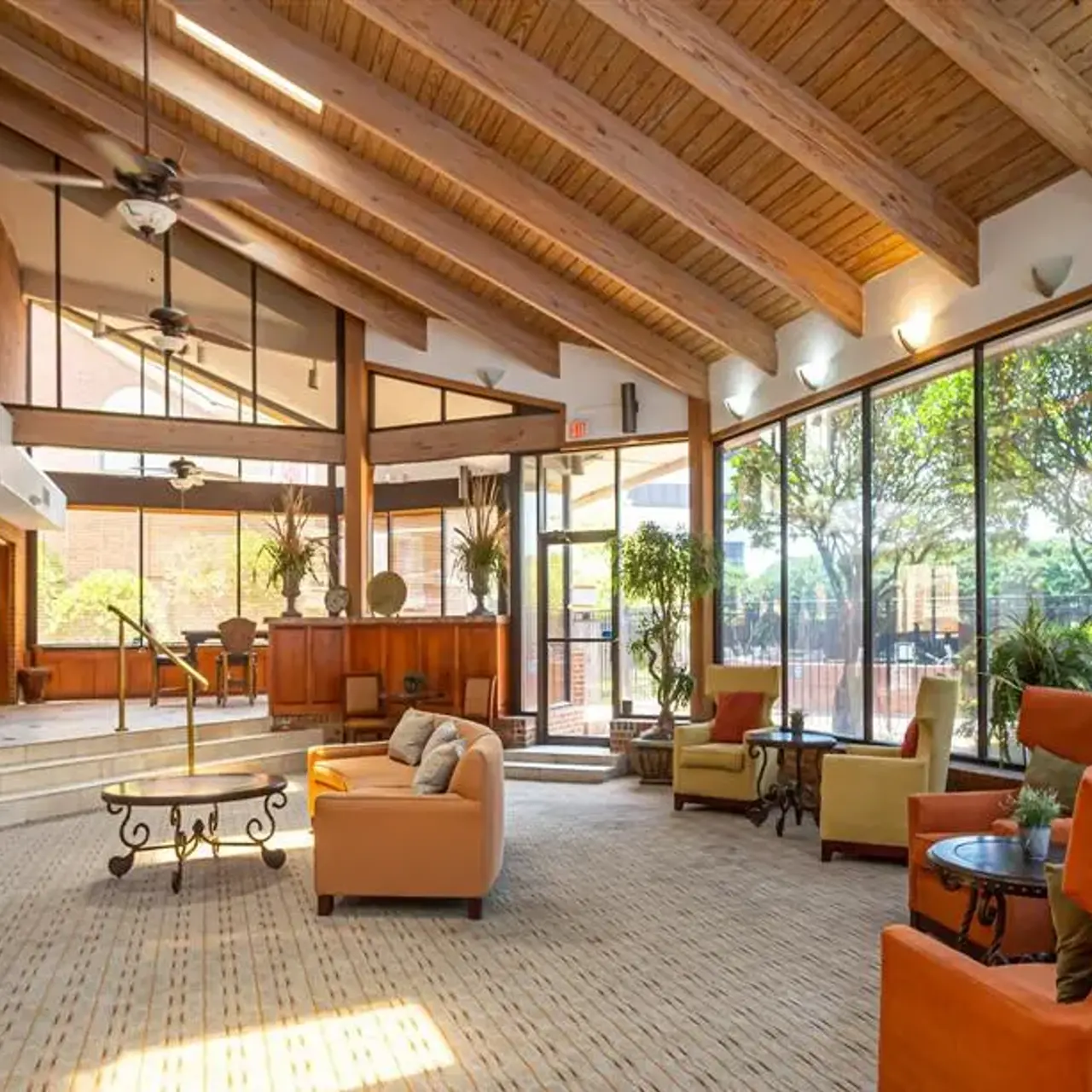 A spacious living area featuring high wooden beams, large windows with a view of greenery, cozy seating arrangements, and a fireplace.