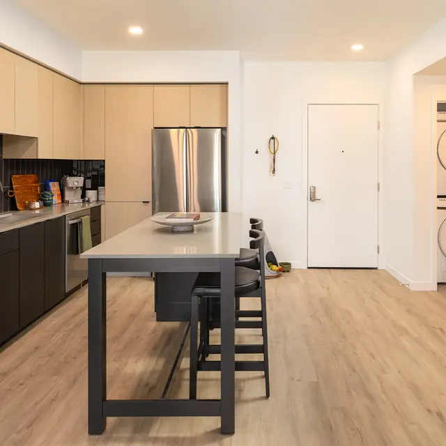 A modern kitchen with stainless steel appliances, a central island, and light wood flooring. The kitchen features a combination of sleek black and beige cabinetry, a large refrigerator, and a laundry area visible in the background.