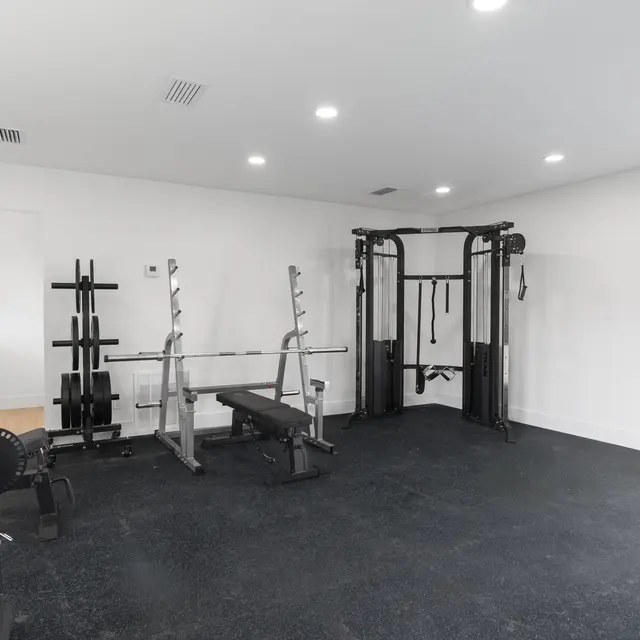 A modern home gym featuring various workout equipment including a multi-functional trainer, a weight bench, and free weights on racks, against a clean white wall and black flooring.