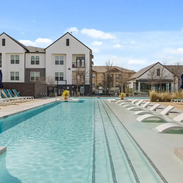 A modern outdoor swimming pool area featuring lounge chairs, umbrellas, and residential buildings in the background.