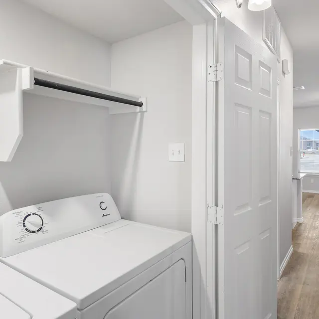 A modern laundry room with a washing machine and dryer, white walls, and a door leading to another room. A shelf above the machines is empty. The flooring is light wood, and there's a glimpse of a well-lit hallway at the end.