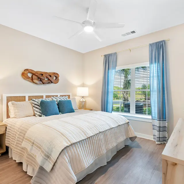 Modern Bedroom Interior A cozy modern bedroom featuring a large bed with light-colored bedding, accent pillows, a wooden headboard, and bedside lamps. There are blue curtains framing a window that lets in natural light, along with a decorative wall hanging. The room has wooden flooring and a light beige color scheme.