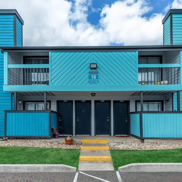 A vibrant blue apartment complex with distinct architectural features, located under a partly cloudy sky. The building has multiple entrances, balconies, and landscaping in the foreground.
