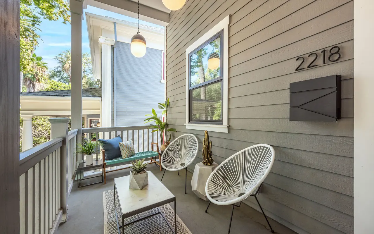 A stylish front porch with two white chairs, a small table, and a decorative piece. The walls are painted grey, and there are hanging light fixtures above. A mailbox is mounted next to the door with the number '2218' displayed.
