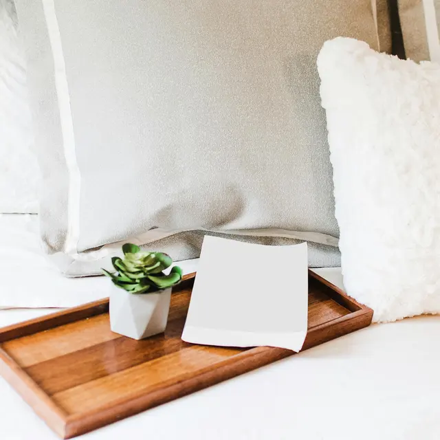 Cozy Bedside Arrangement A cozy bedside setup featuring a wooden tray with a small white pot containing a succulent, a blank sheet of paper, and a fluffy white pillow on a bed with light gray pillows.