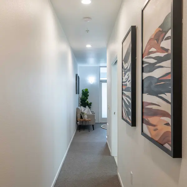 A well-lit hallway featuring two framed artworks on the wall, with a small seating area visible at the end and greenery in a pot.