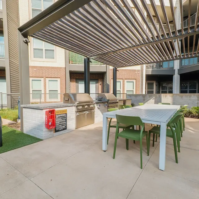 An outdoor grilling area featuring a large dining table with green chairs under a pergola, a fully equipped grill station, and a large umbrella on the side.