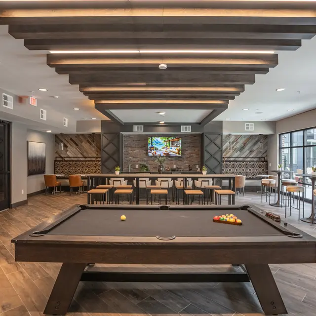 A modern game room featuring a pool table and a lounge area with tables and chairs, illuminated by stylish ceiling lights. There is a large wall TV and decorative elements in the background.