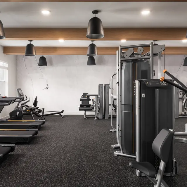 A modern gym interior featuring treadmills, exercise machines, and black rubber flooring, illuminated by industrial-style pendant lights.