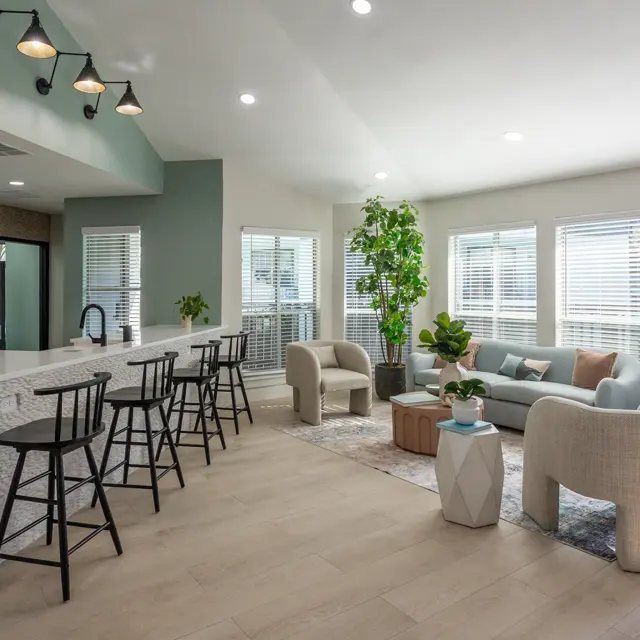 A modern living space featuring a bar counter with stools, a comfortable seating area with a couch and armchairs, large windows providing natural light, and indoor plants.