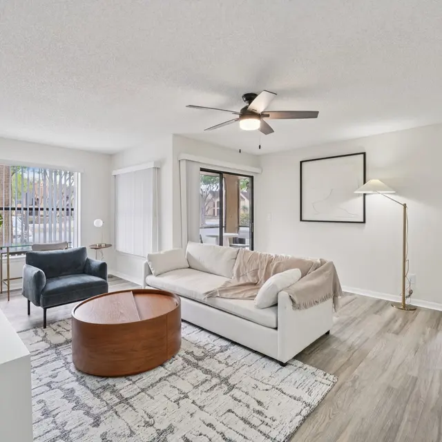 A modern living room featuring a white sofa, round wooden coffee table, and a blue accent chair. There is a large window with blinds, a potted plant, and light-colored wooden flooring.