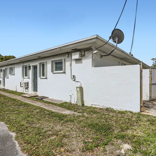 Side view of a small apartment complex with white walls and a fenced entrance. The area is grassy with gravel paths, and there are utility poles in the background.