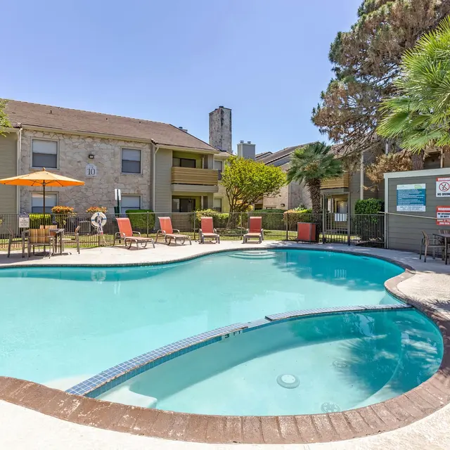 A bright, inviting swimming pool located in an apartment complex courtyard, surrounded by lounge chairs and lush landscaping under clear blue skies.