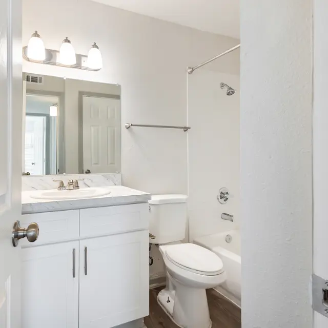 A modern bathroom featuring a white vanity with a marble countertop, a bathtub with a shower, and a large mirror above the sink. The walls are painted in a light color, and the floor has a wood-like appearance.