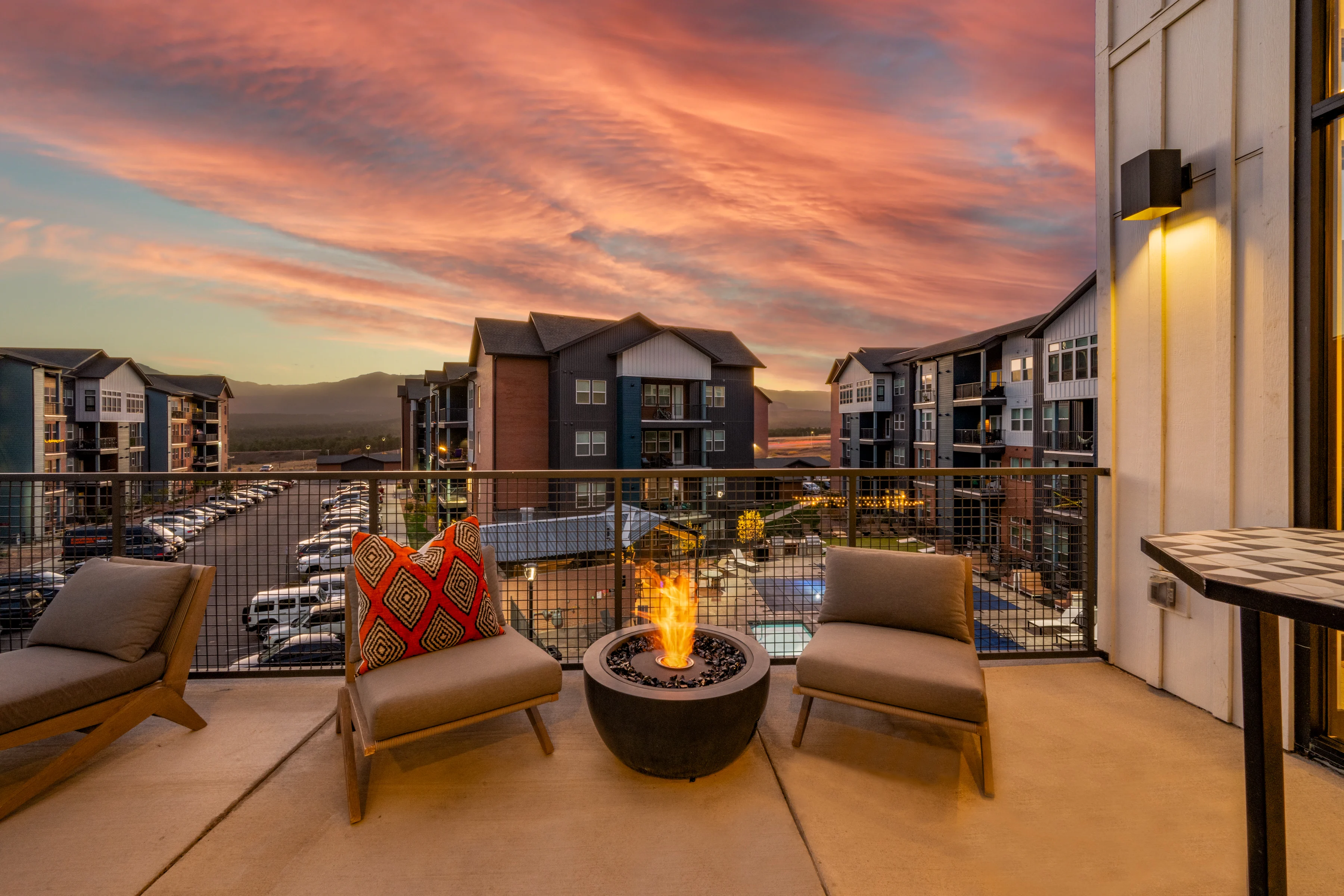 Luxury apartment balcony with chairs and a fire pit overlooking a sunset sky and parking lot.