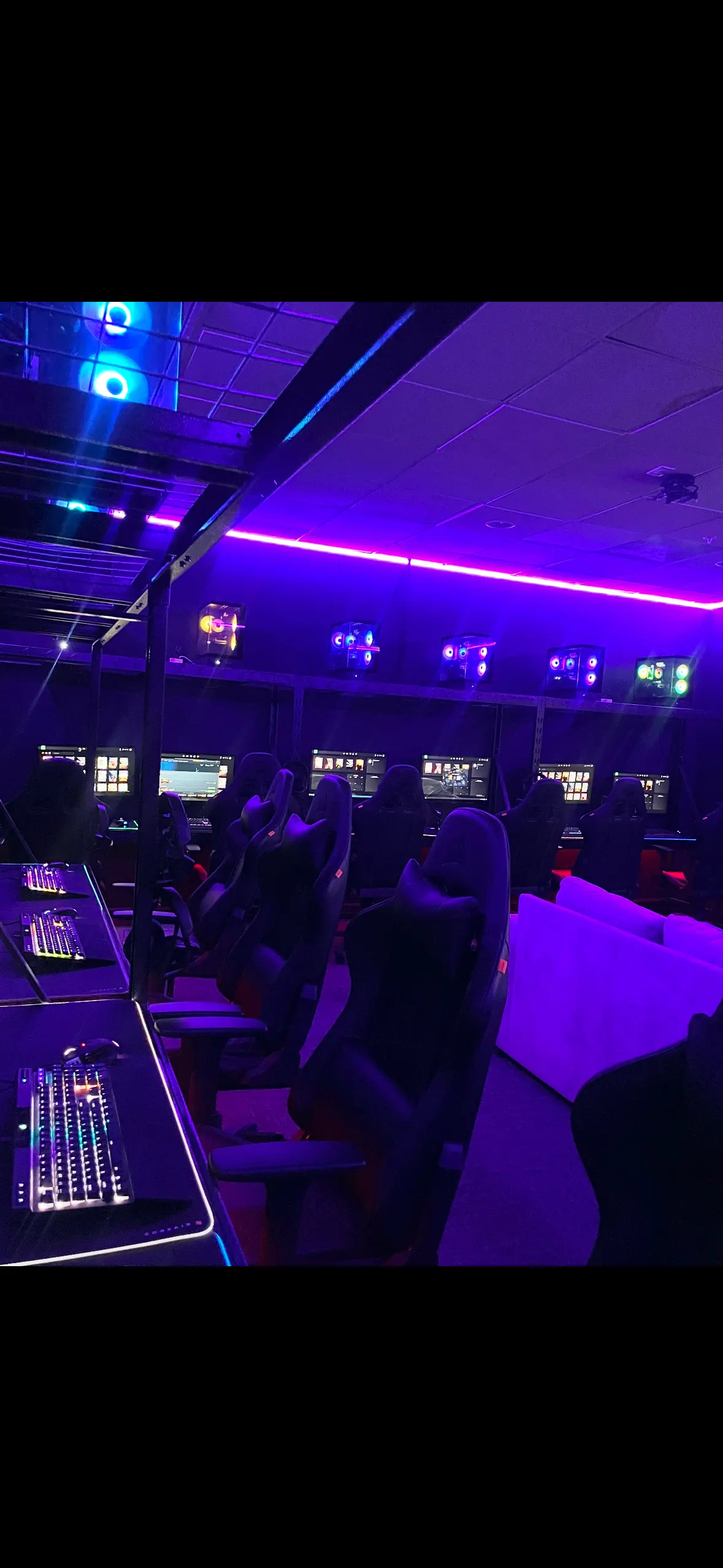 Modern Gaming Lounge Interior An interior view of a gaming space featuring multiple high-tech gaming chairs and computer setups. The room is illuminated with purple LED lights, creating an immersive atmosphere. Monitors are placed on desks in front of each chair, and additional seating is visible against a back wall.