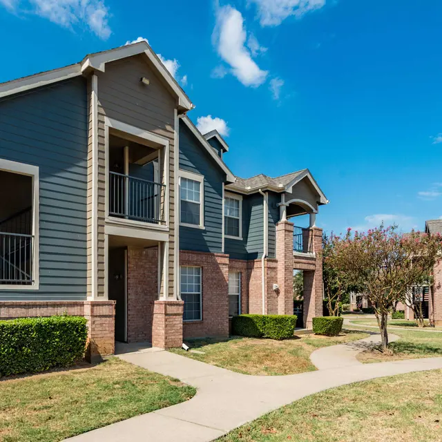 Exterior view of a modern apartment complex featuring two buildings, green landscaping, and a blue sky.