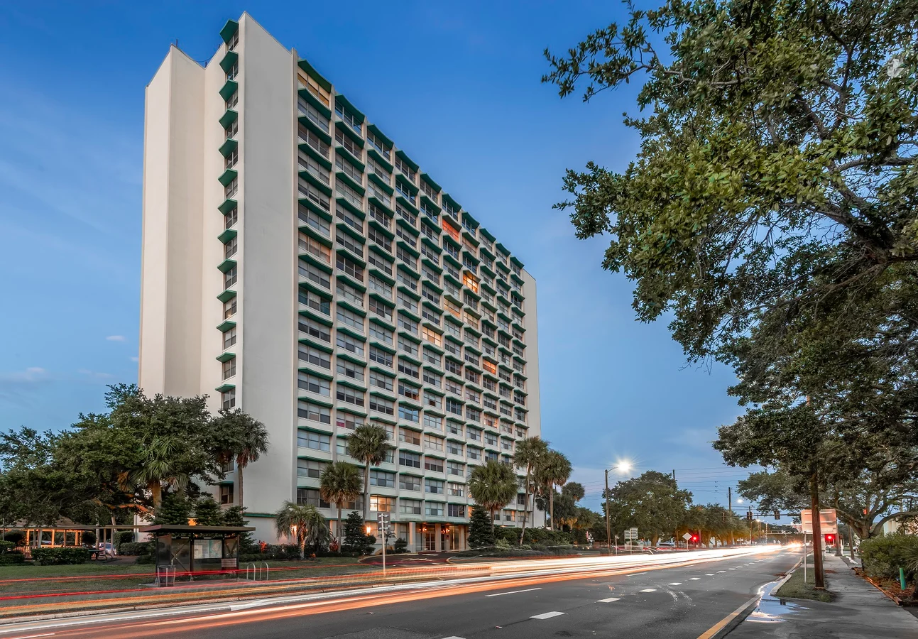 A modern high-rise apartment building with multiple floors and large windows, set against a dusk sky. The building is surrounded by palm trees and street lights, with light trails from passing cars in the foreground.