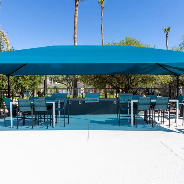 A large blue shade structure with seating underneath, surrounded by palm trees and greenery in an outdoor area. The ground is white concrete, and there is a fenced area in the background.