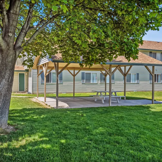 Outdoor Pavilion in Green Space A grassy area featuring a wooden pavilion with a concrete floor. The pavilion is shaded by trees and has picnic tables underneath. In the background, there are residential buildings.