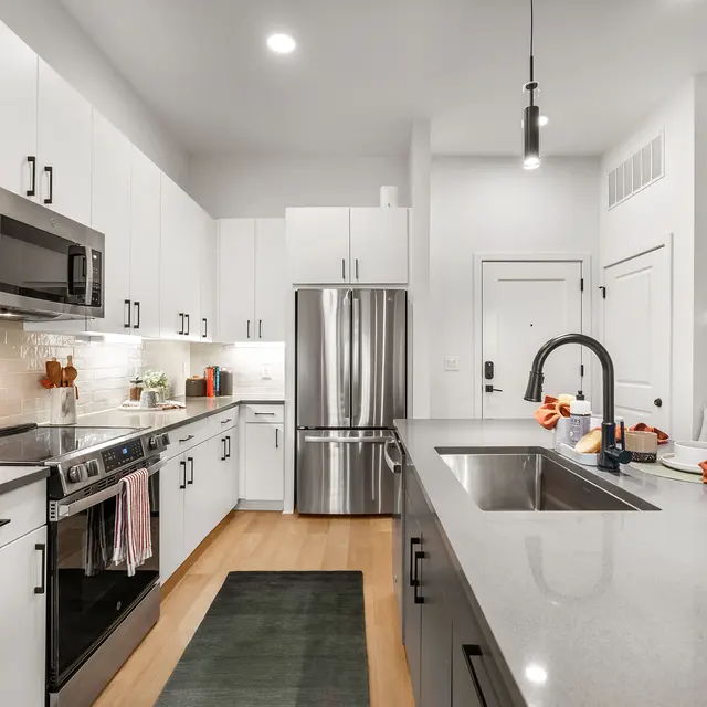 A contemporary kitchen featuring white cabinetry, stainless steel appliances, and a large island with a sink. The kitchen has a bright, open layout with warm wood flooring and stylish decorative elements.