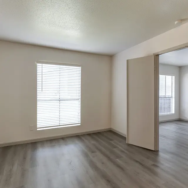 An empty room with light-colored walls and flooring. One window with blinds is visible, allowing natural light to enter. There is an open doorway leading to another room with similar flooring.