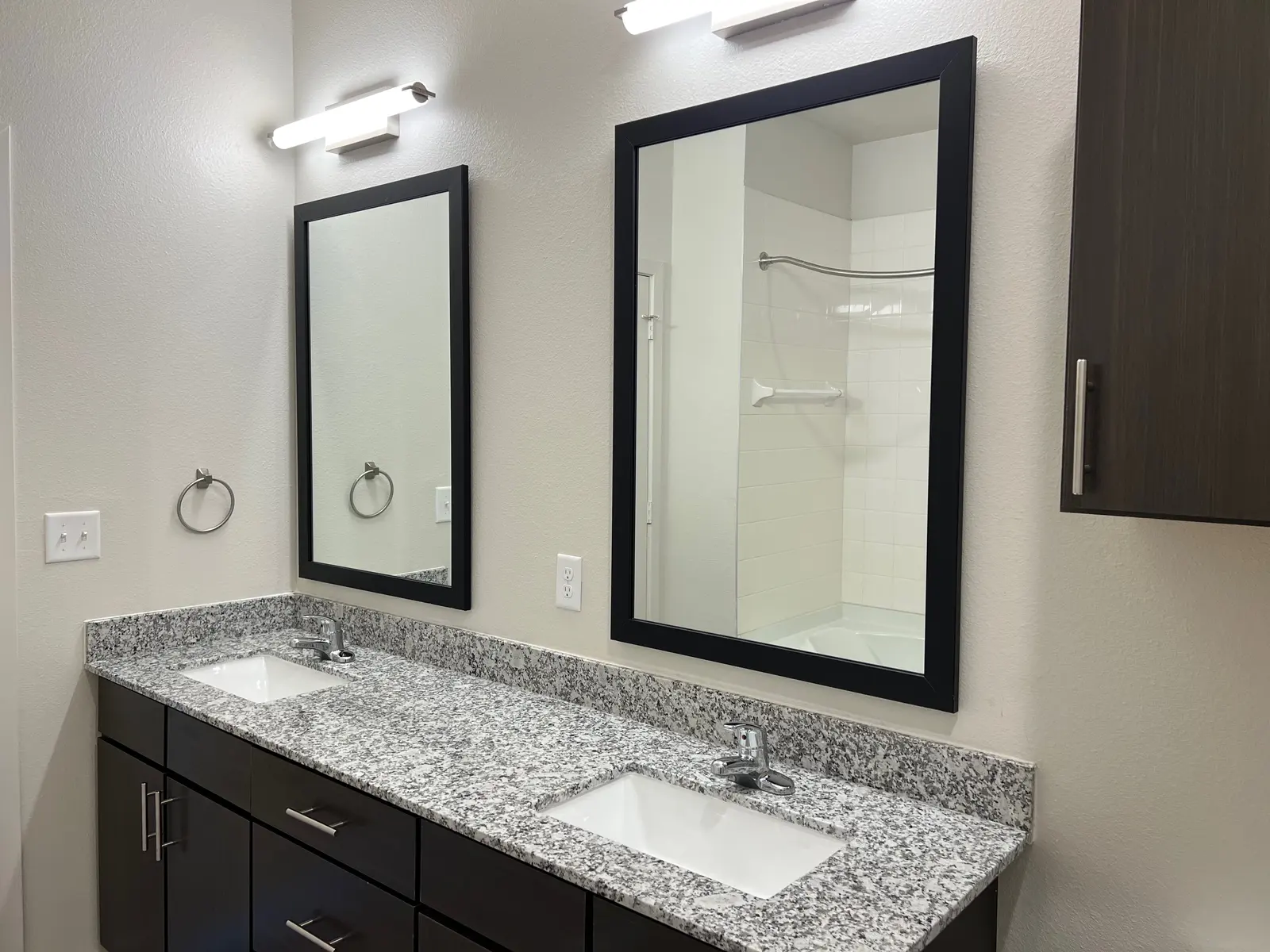 A modern bathroom featuring a double vanity with two sinks, dark wood cabinetry, and two framed mirrors mounted on the wall, illuminated by wall sconces. A shower area is visible in the background.