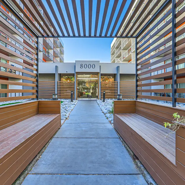 A modern apartment building entrance featuring wooden slats and concrete pathways leading to the front door.