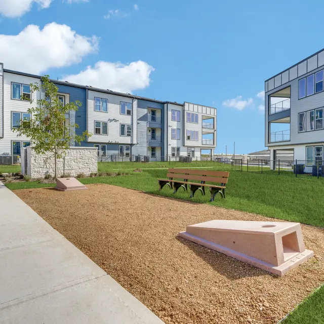 A modern apartment complex with a green lawn area, benches, and a gravel surface. Featuring contemporary architecture with large windows.