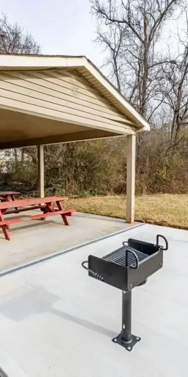 A covered picnic area with red picnic tables and built-in grills in a grassy park setting.