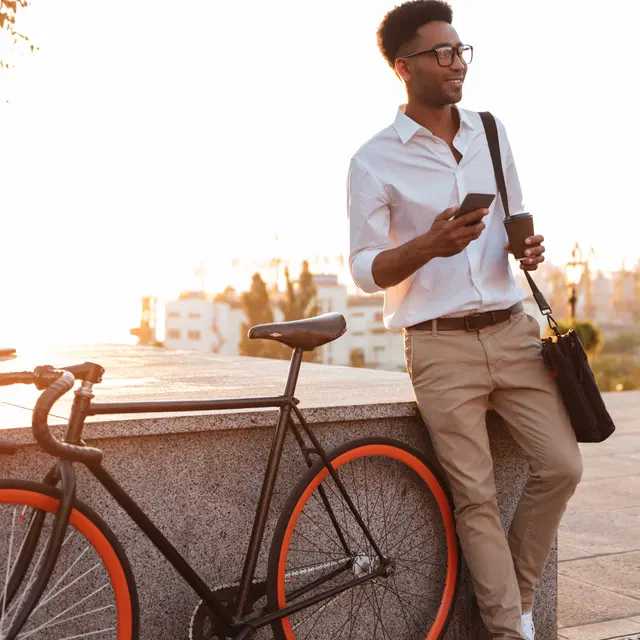 A man in a white shirt and beige pants standing next to a black bicycle with orange accents, holding a smartphone, while a warm sunset illuminates the background.