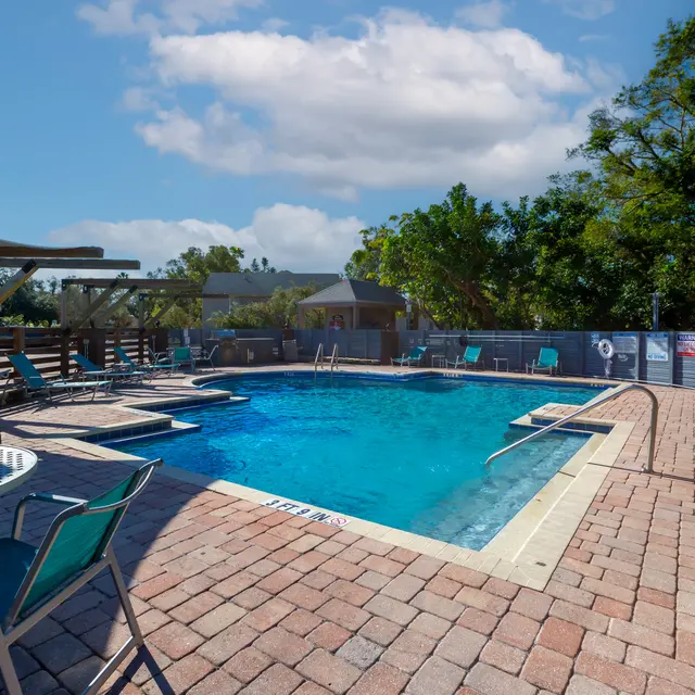 A serene pool area with a blue swimming pool surrounded by brick pavement, poolside chairs, and shaded seating nearby.