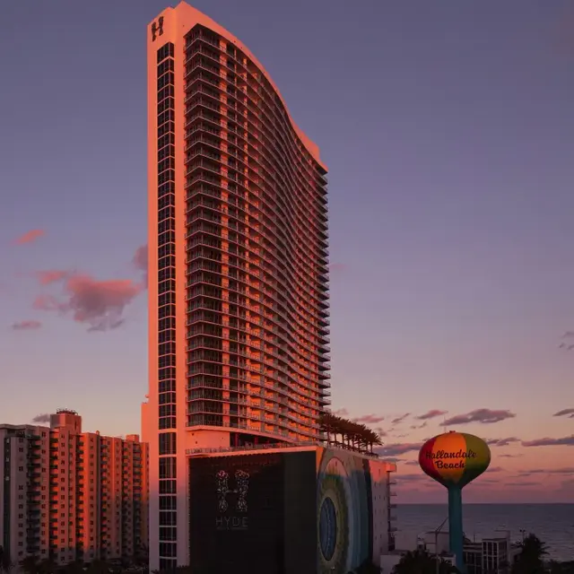 Hyde Hotel at Sunset A tall modern building with a curved top, featuring many windows, set against a colorful sunset sky. In the foreground, a round sign displays 'Hollywood Beach'.