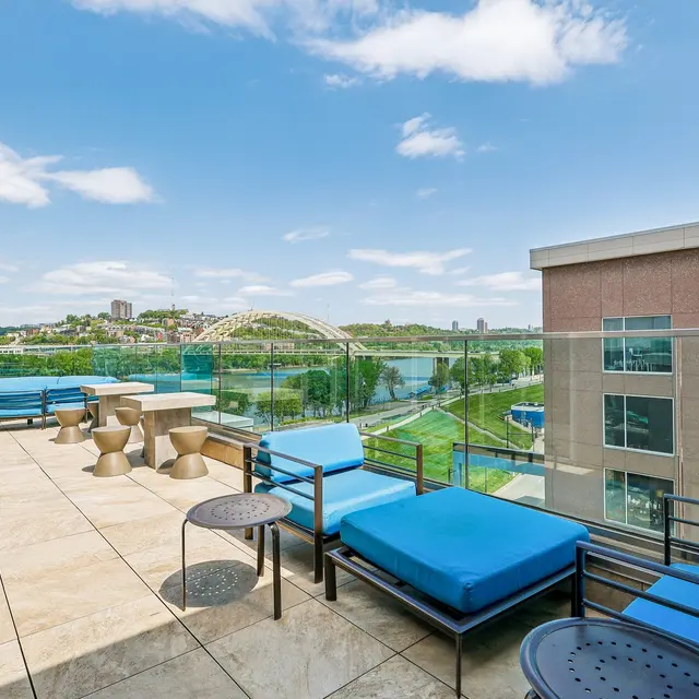 A spacious rooftop terrace with modern furniture, featuring blue lounge chairs and small tables, overlooking a green landscape and a body of water under a bright blue sky with scattered clouds.