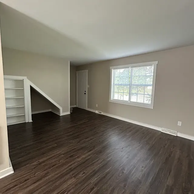 A spacious empty living room with wooden flooring and a window allowing natural light. There is a staircase on the left leading to an upper level, and a door on the right side of the image.