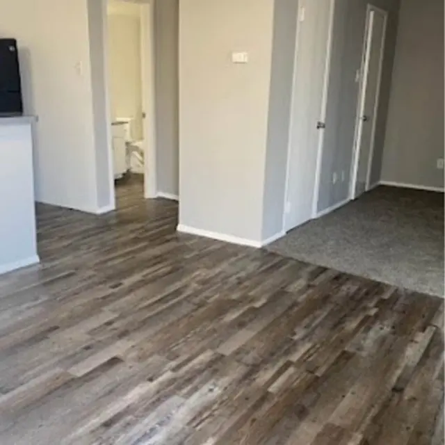 A view of an apartment interior featuring a wooden floor and gray walls. The layout shows a segment of the living area leading to doorways and a carpeted section in the background.