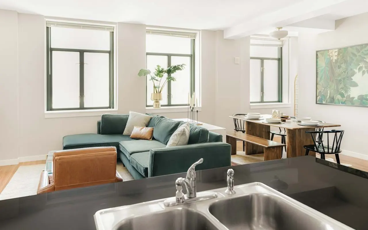 A modern living room featuring a green sofa, a wooden dining table, and large windows that bring in natural light.