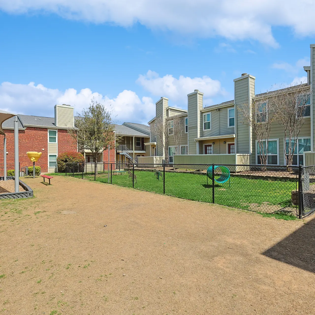 A playground area with a red slide, grass, and several buildings in the background under a blue sky with fluffy clouds.