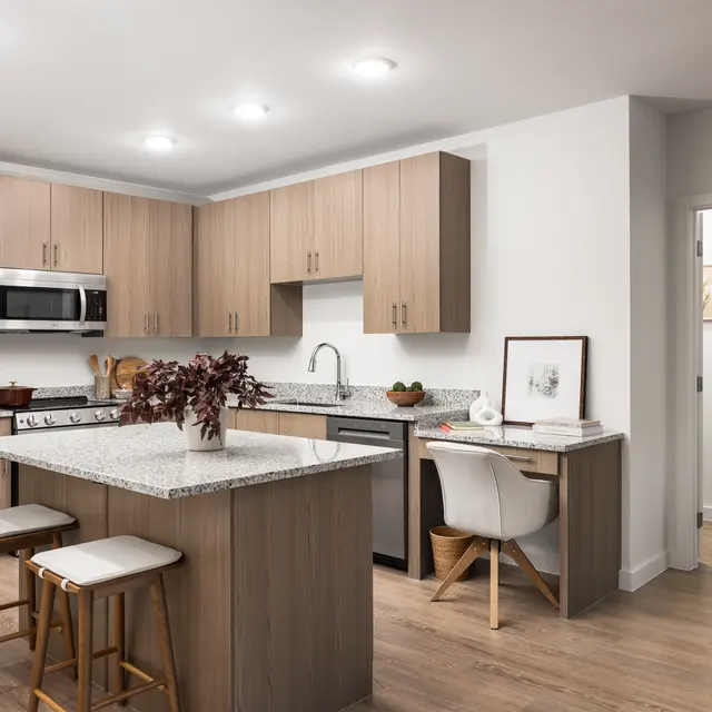 A modern kitchen with wooden cabinetry, a granite island countertop, and bar stools. Appliances include a microwave and stainless steel refrigerator. Natural light illuminates the space, enhancing the neutral color palette. A small dining area is visible with a white chair and decorative items on the countertop.