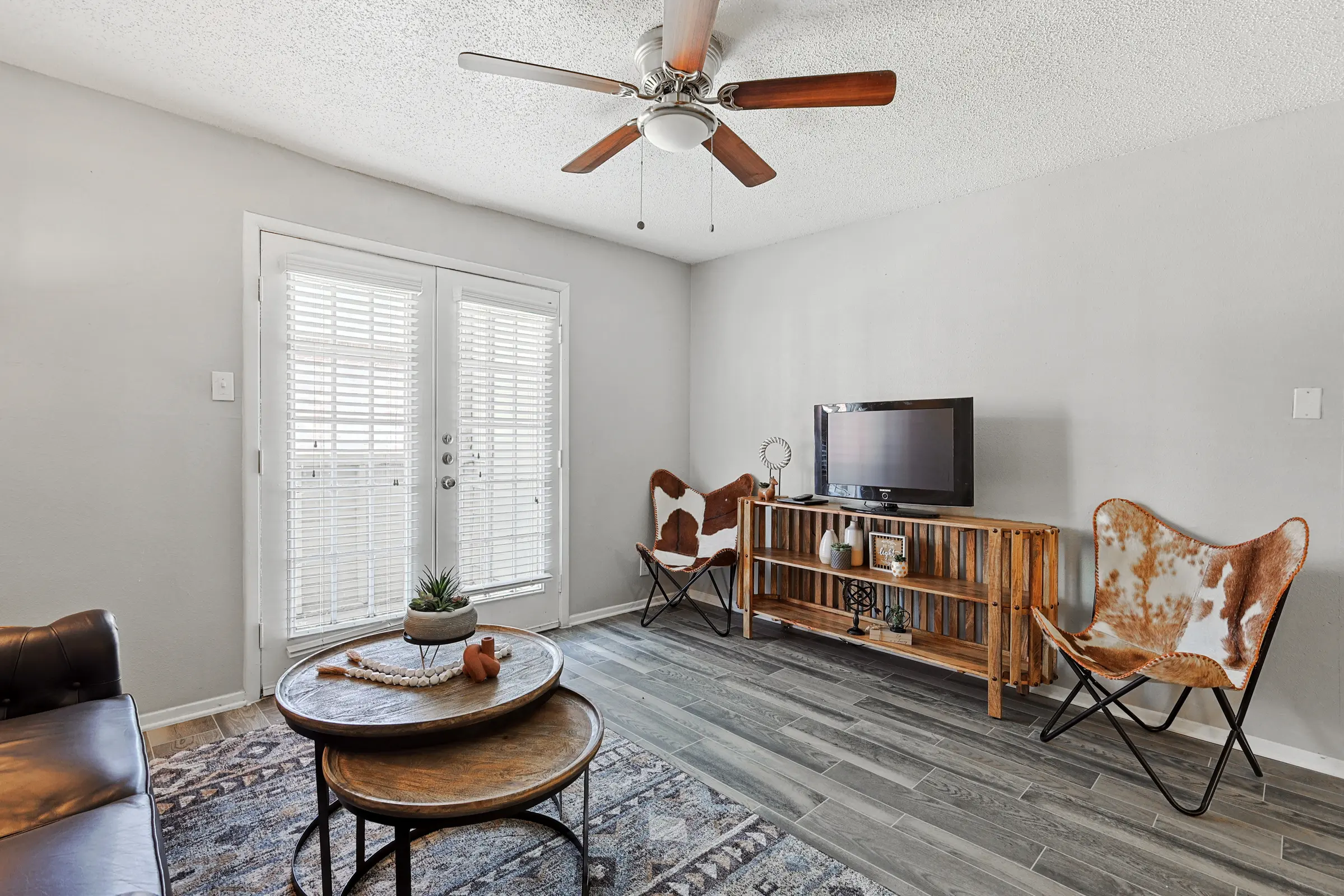 A cozy living room with a ceiling fan, a brown leather sofa, two butterfly chairs, a wooden media console, and coffee tables. There are sliding doors with white shutters letting in natural light.