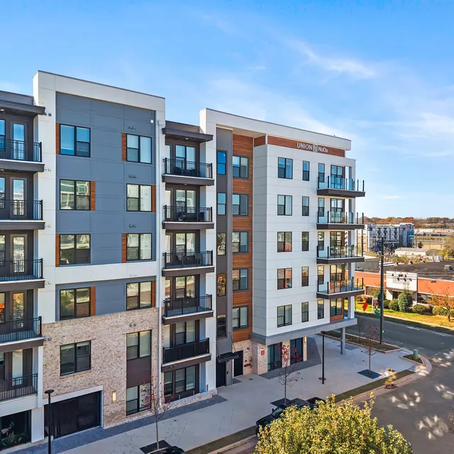 Modern apartment building with multiple floors, featuring balconies and a mix of materials including brick and stucco. Clear sky in the background.