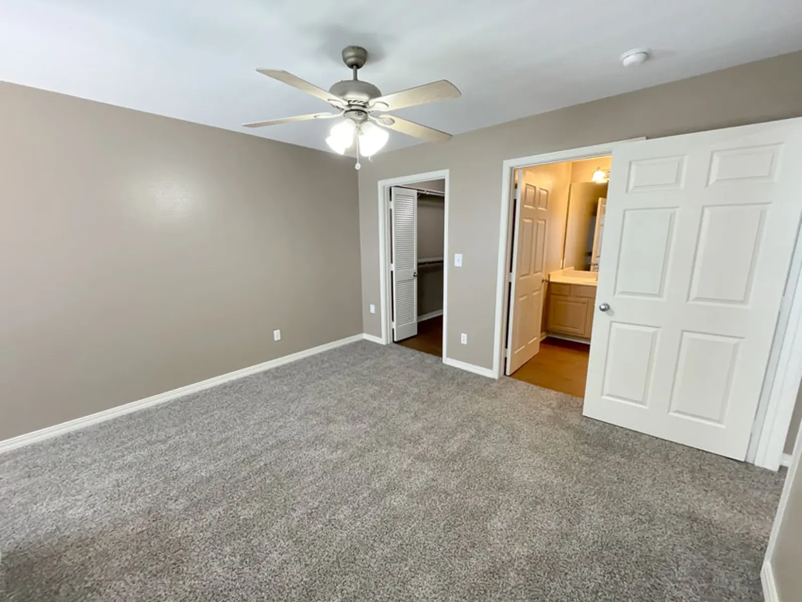 A spacious bedroom featuring light brown walls, plush carpet, and a ceiling fan. There are two doorways leading to a closet and a bathroom both framed in white.