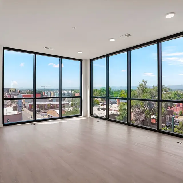 A spacious, modern apartment with large floor-to-ceiling windows offering a view of the cityscape and greenery outside. The room features light hardwood flooring and is empty, allowing for various furnishing possibilities.