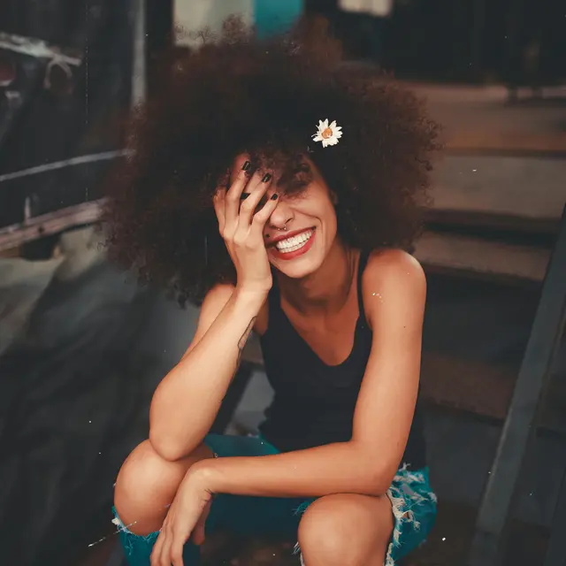 A young woman with curly hair is sitting on a staircase, smiling while covering part of her face with her hand. She wears a black tank top and ripped blue jeans, with a daisy flower in her hair.