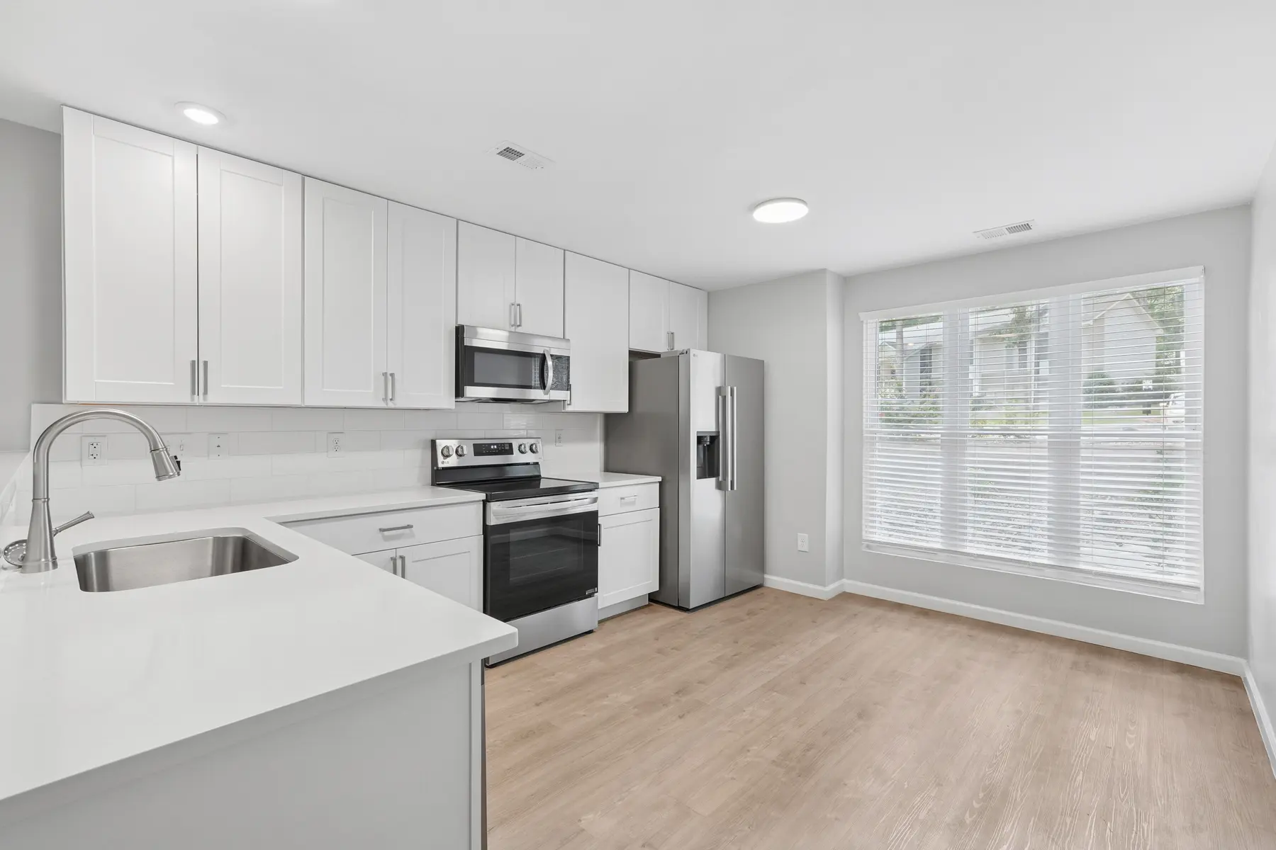 A modern kitchen featuring white cabinets, stainless steel appliances, and a large window with blinds allowing natural light in. The flooring is a light wood tone, and the countertop is white.