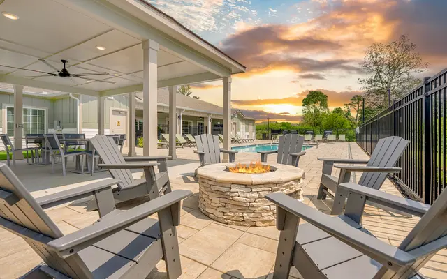 An outdoor seating area with gray Adirondack chairs around a stone fire pit, near a swimming pool, under a covered patio with a beautiful sunset in the background.
