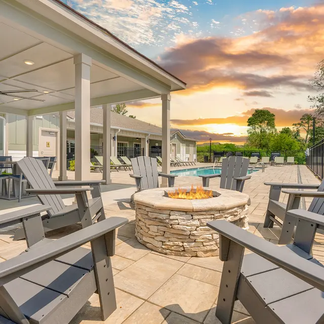 An outdoor seating area with gray Adirondack chairs around a stone fire pit, near a swimming pool, under a covered patio with a beautiful sunset in the background.