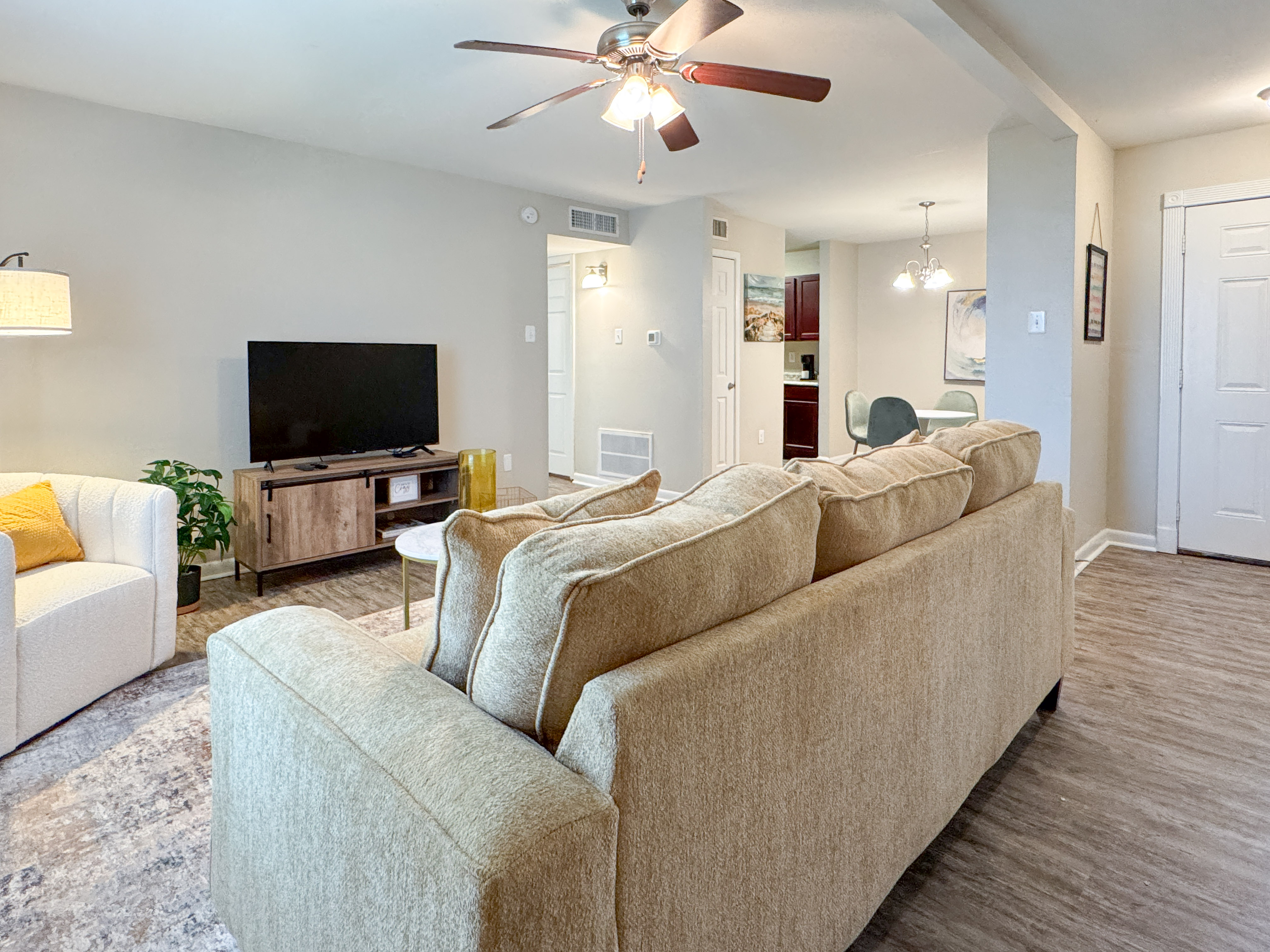 A cozy living room featuring a light-colored sofa, a single white armchair with a yellow cushion, a TV unit with a flat-screen TV, and a small dining area visible in the background. The room has a ceiling fan and a beige rug on the wooden floor.