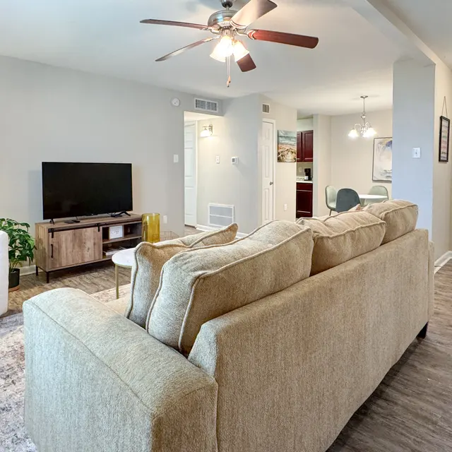 A cozy living room featuring a light-colored sofa, a single white armchair with a yellow cushion, a TV unit with a flat-screen TV, and a small dining area visible in the background. The room has a ceiling fan and a beige rug on the wooden floor.